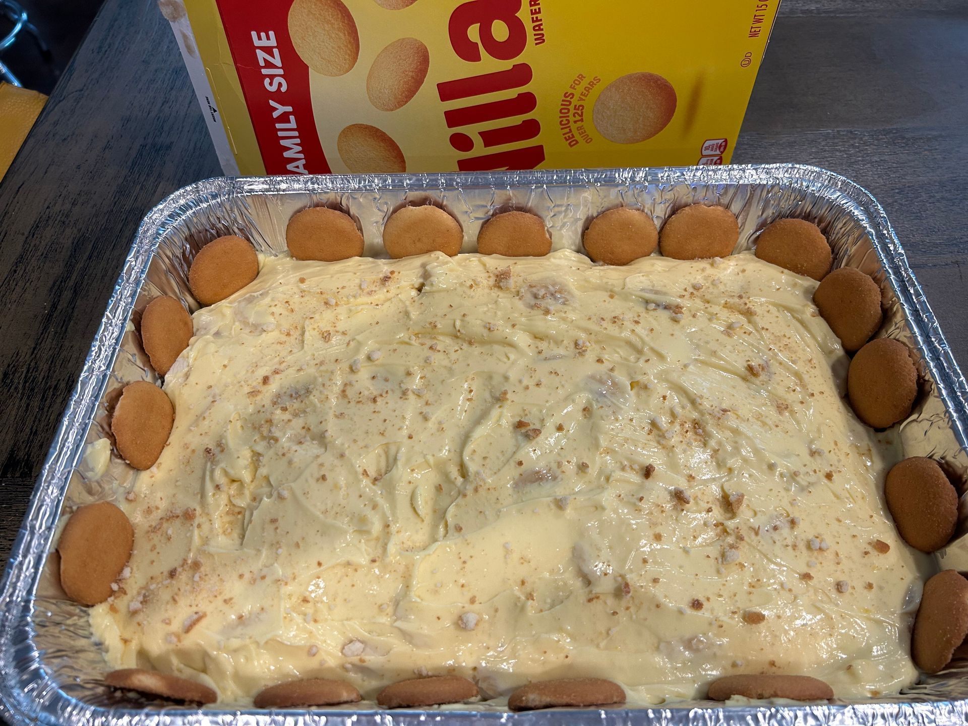 Banana pudding in a foil pan, with cookies around the edge, and a box of cookies behind it.