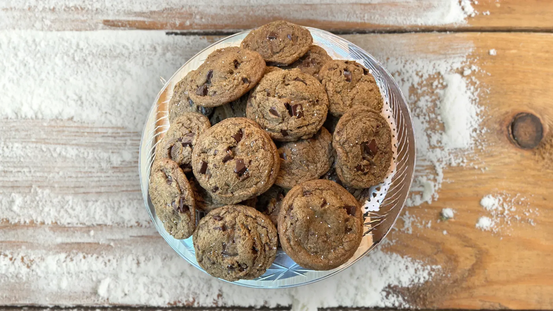 Chocolate chip cookies on a plate, surrounded by flour on a wooden surface.