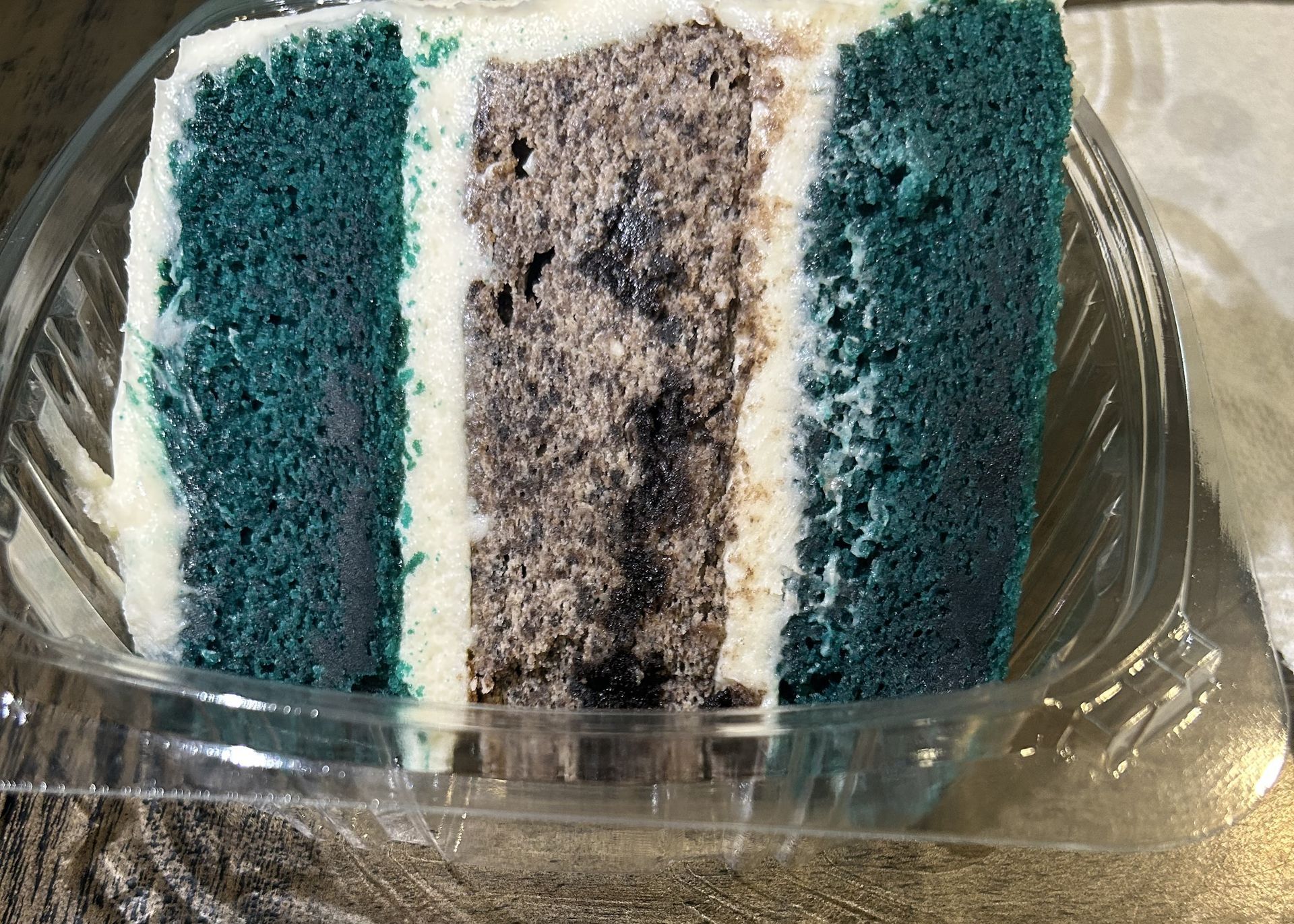 Slice of cake: teal outer layers, brown interior, white frosting in a clear container.