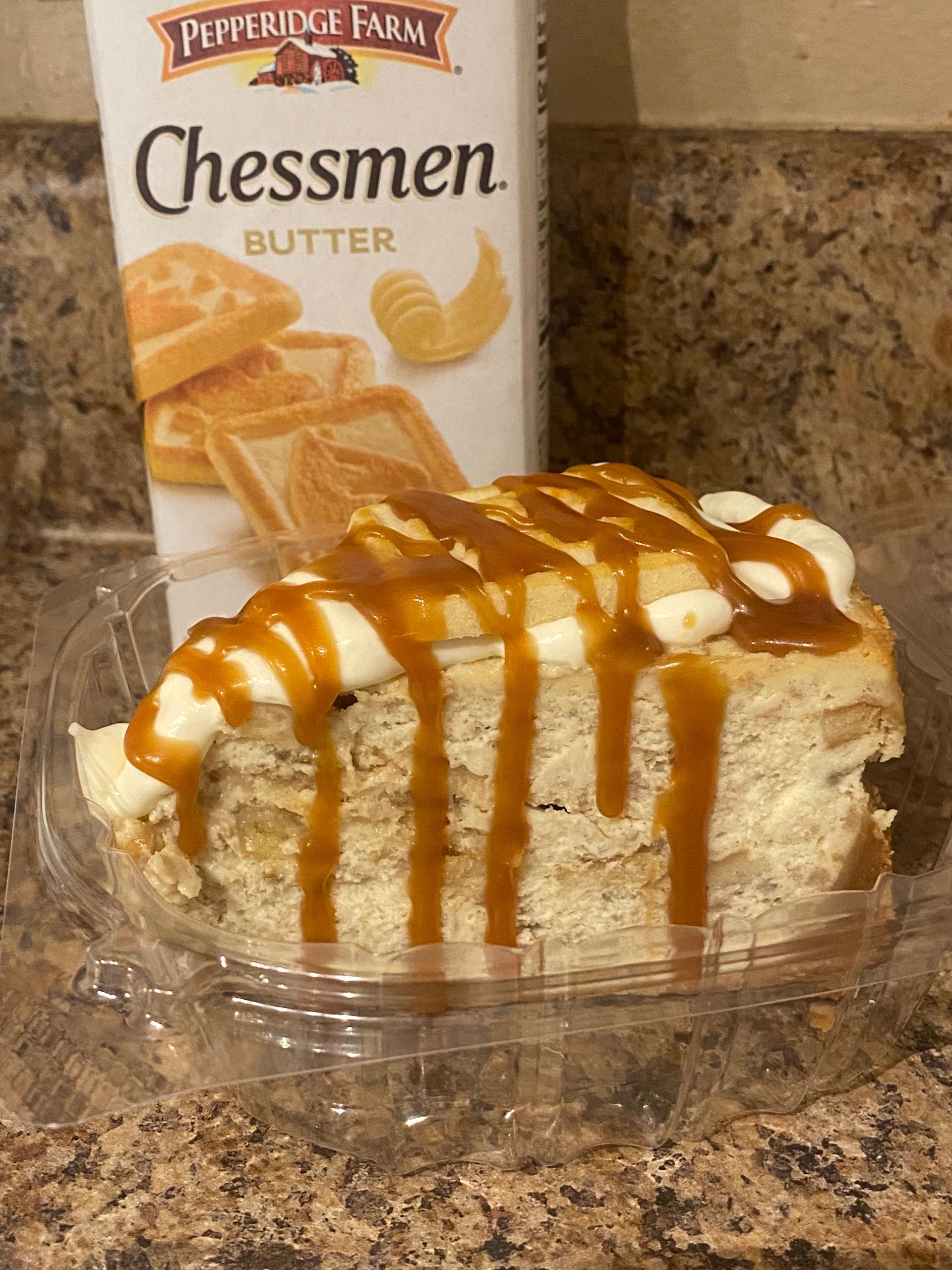 Slice of cake with caramel drizzle in a clear plastic container next to a box of Pepperidge Farm Chessmen cookies.