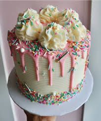 Cake with white frosting, pink drip, and colorful sprinkles, topped with buttercream swirls.