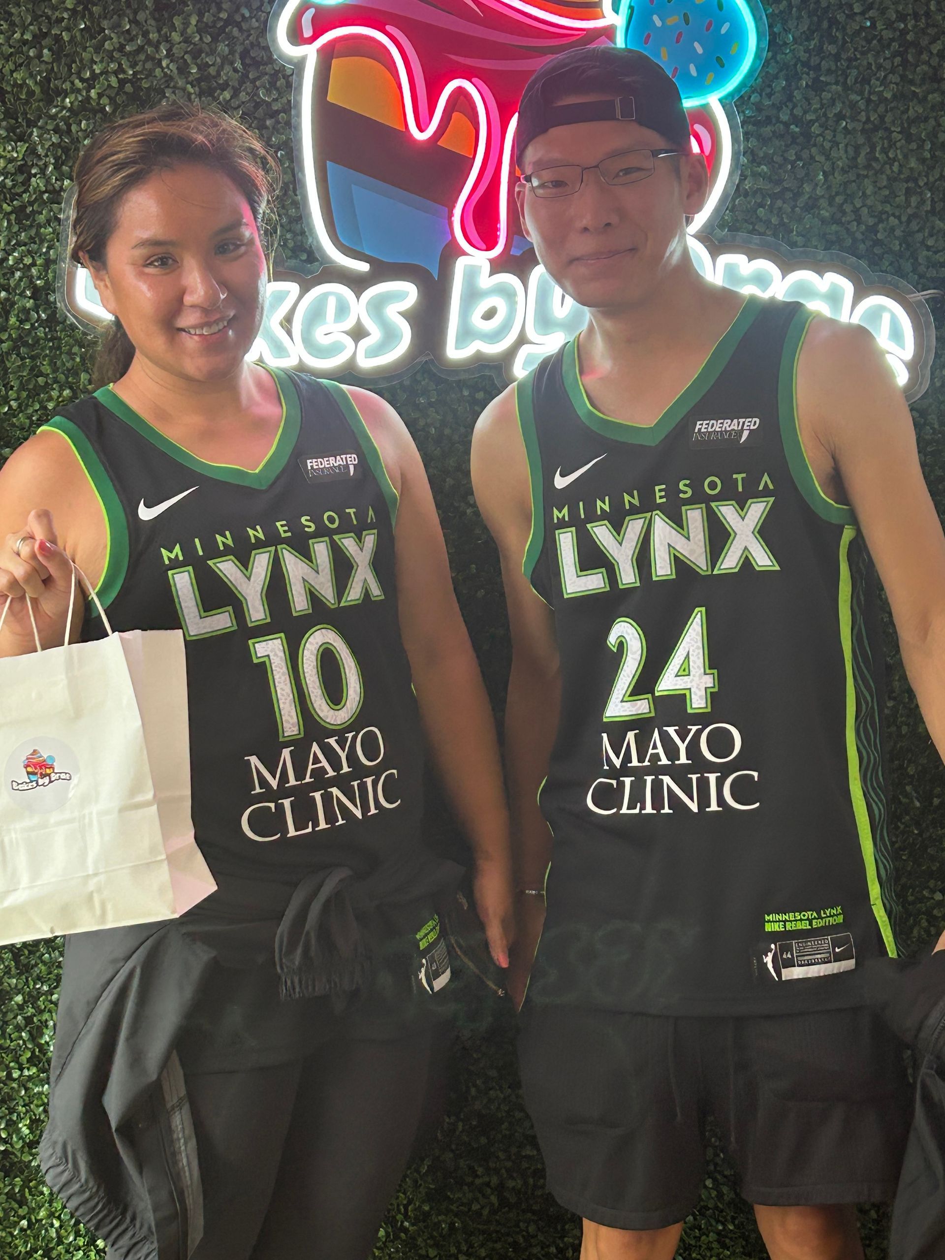 Two people wearing Minnesota Lynx jerseys pose in front of a bakery sign.