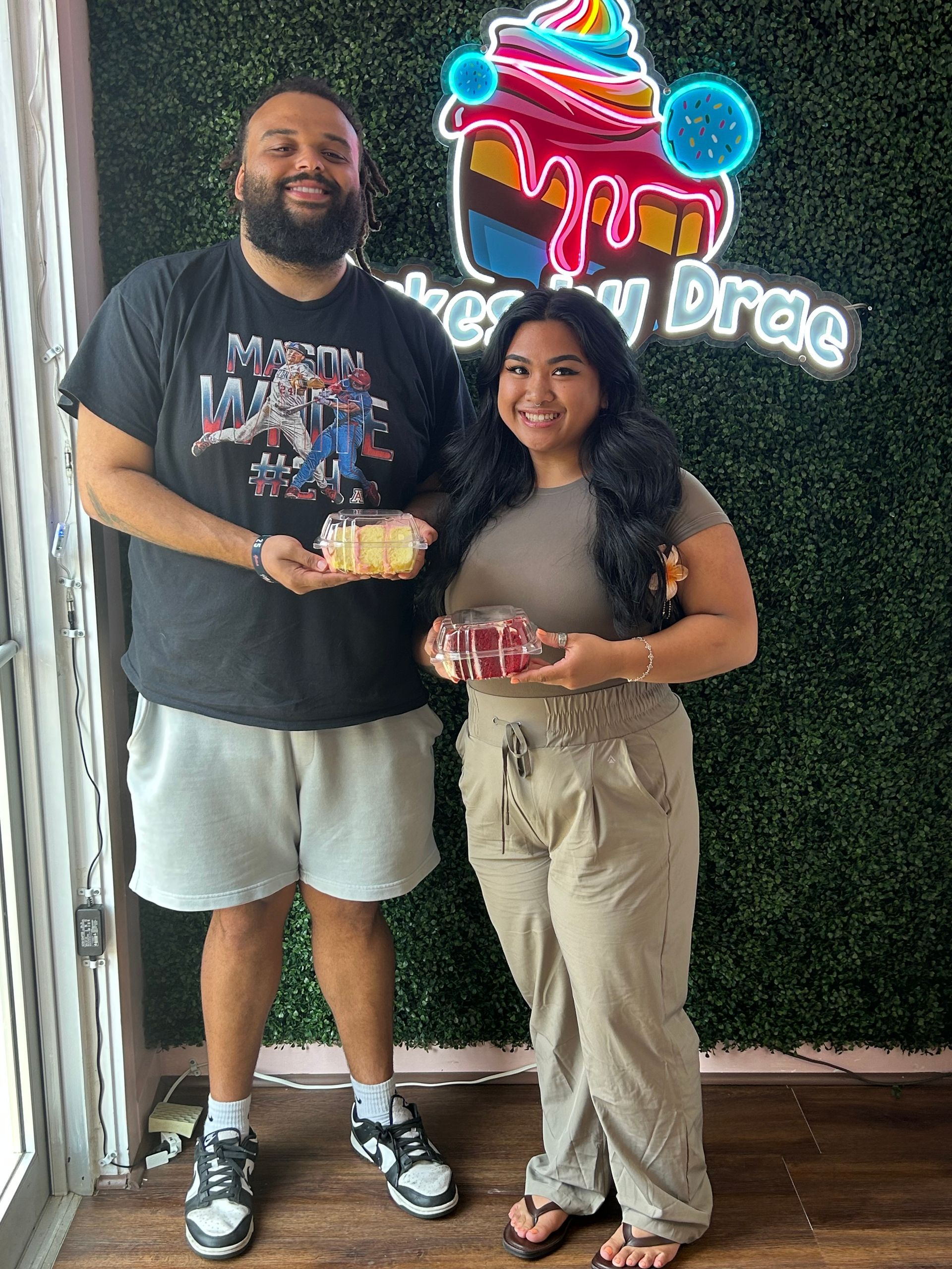 Man and woman holding cake and gift in front of a bakery sign.