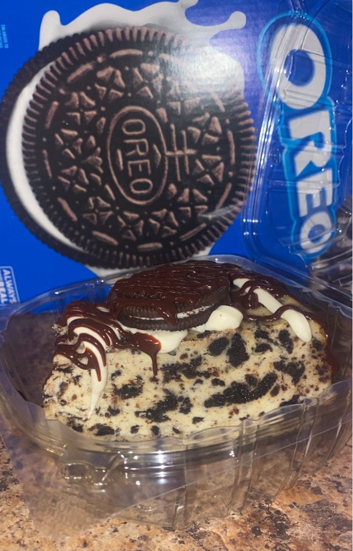 Oreo cheesecake slice in a clear container with chocolate drizzle, next to a blue Oreo cookie package.