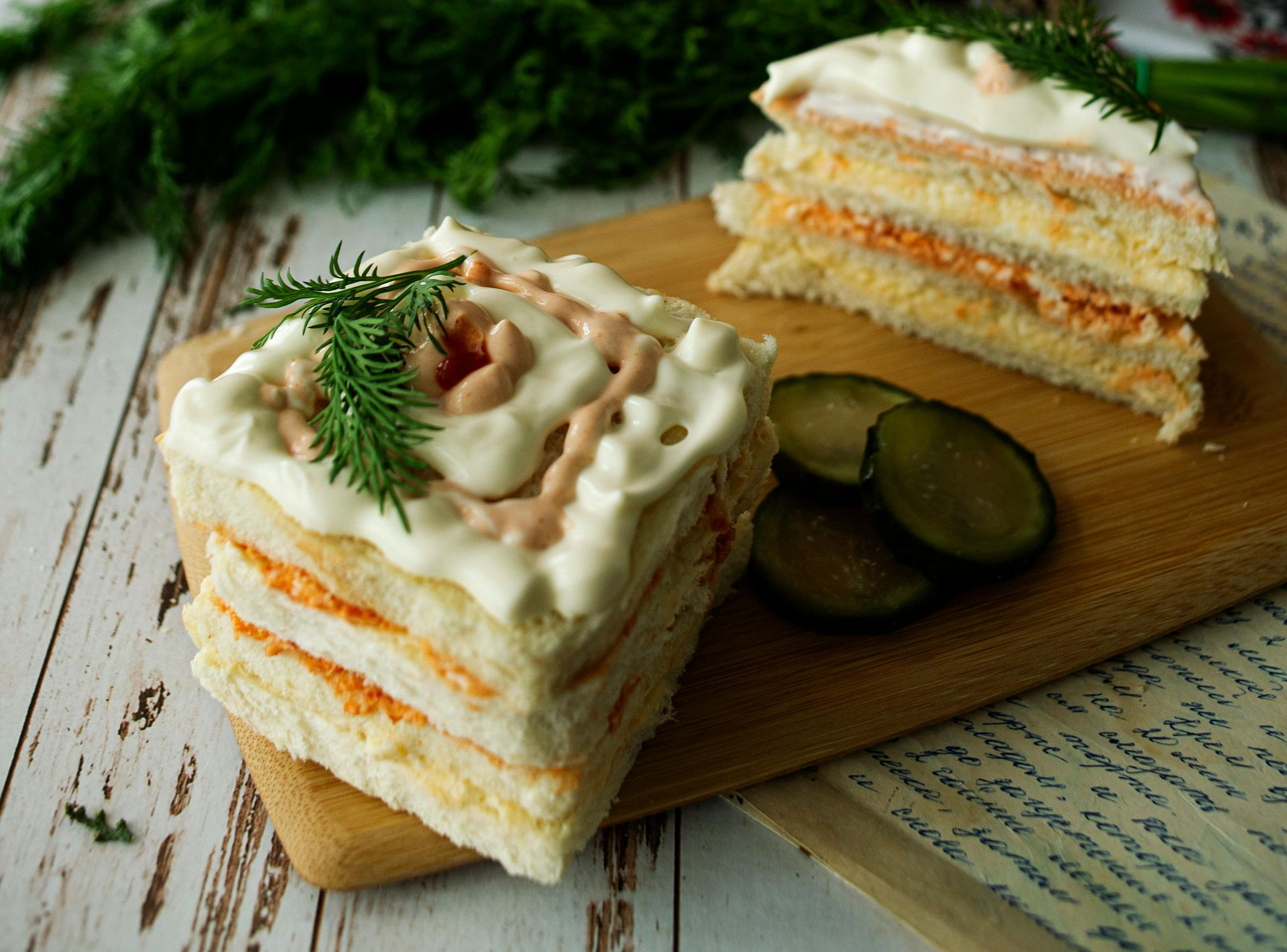 Sandwich cake slices with orange filling, white frosting, dill garnish, and pickles on a wooden board.