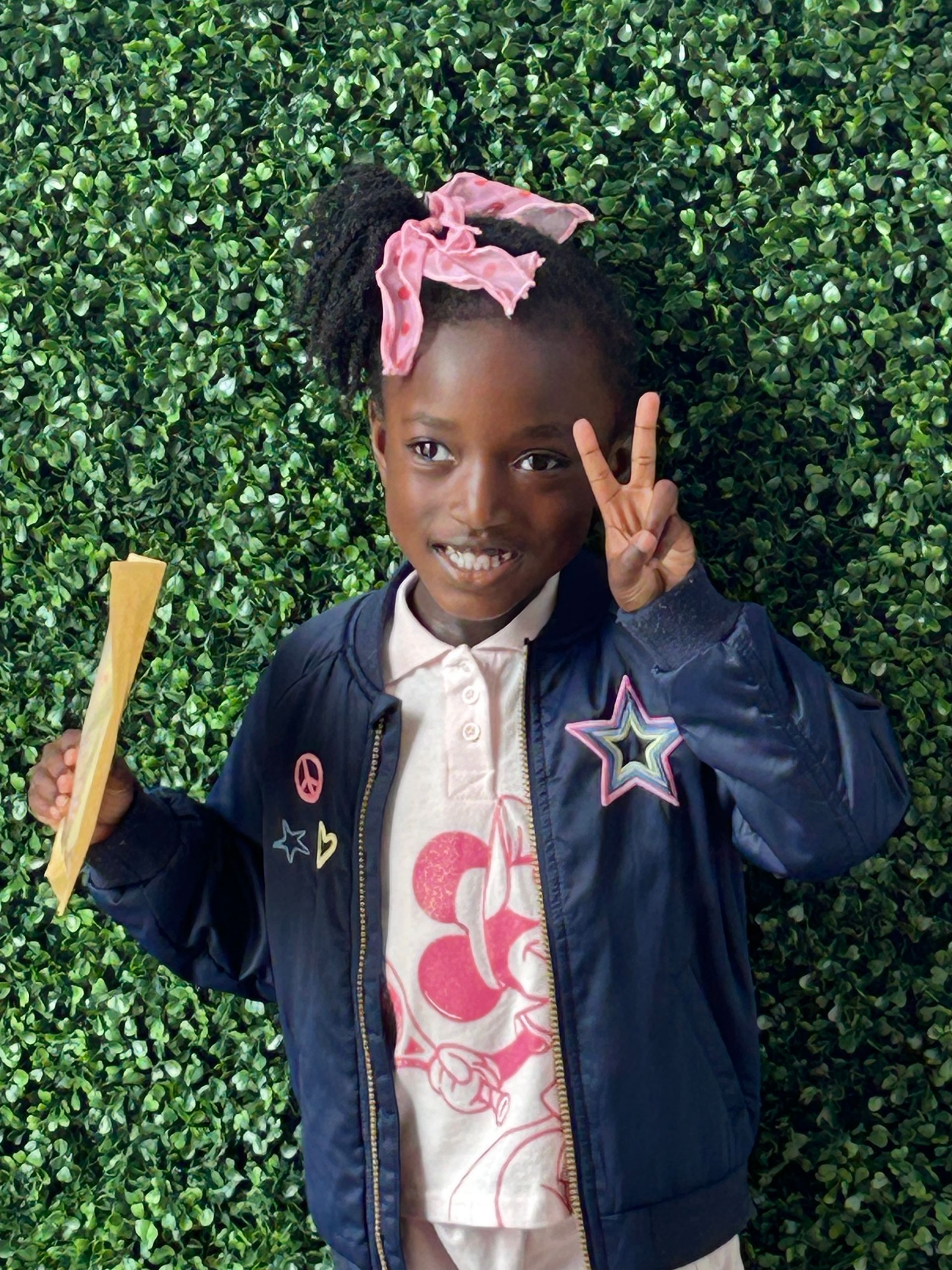 Girl with a high ponytail wearing a blue jacket and pink shirt, making a peace sign in front of a green wall.
