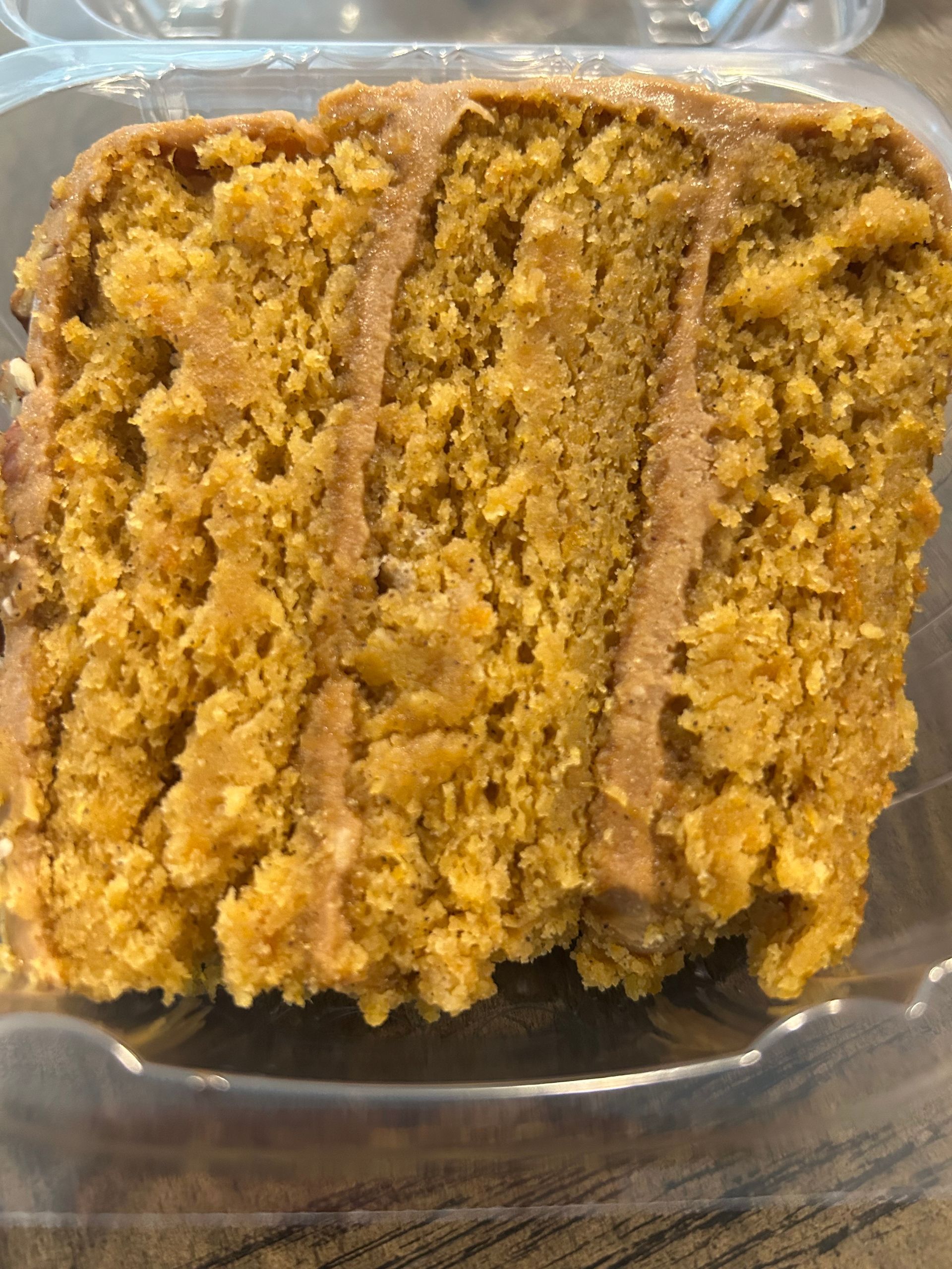 Slice of pumpkin spice cake with brown icing in a clear plastic container.