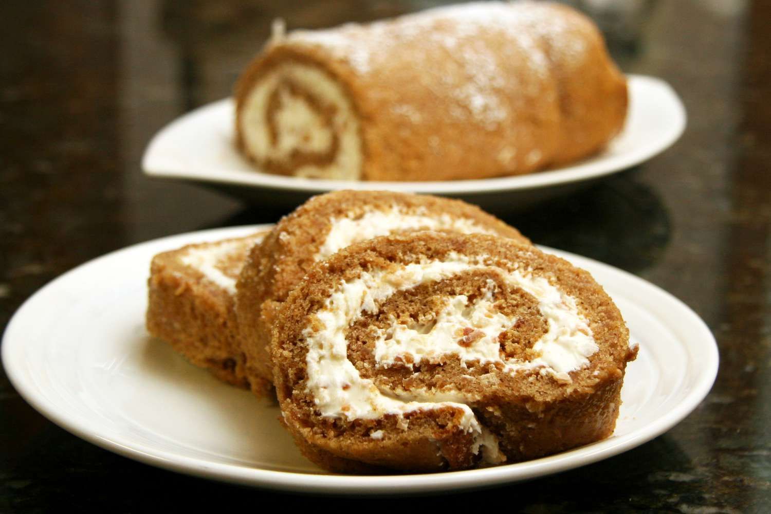 Chocolate swiss roll cake slices on a plate, another roll in the background, dusted with powdered sugar.