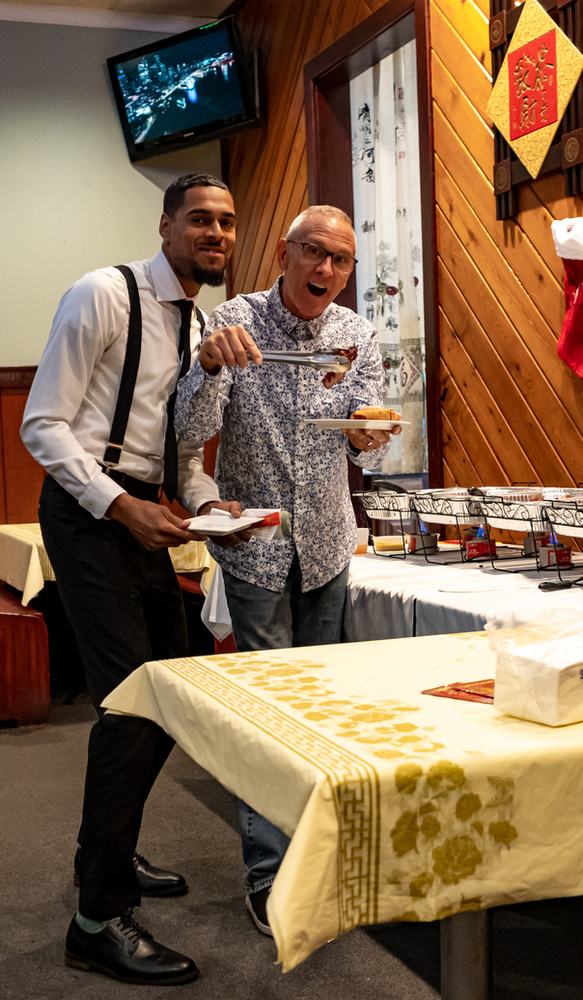 Two men are standing next to each other at a table holding plates of food.