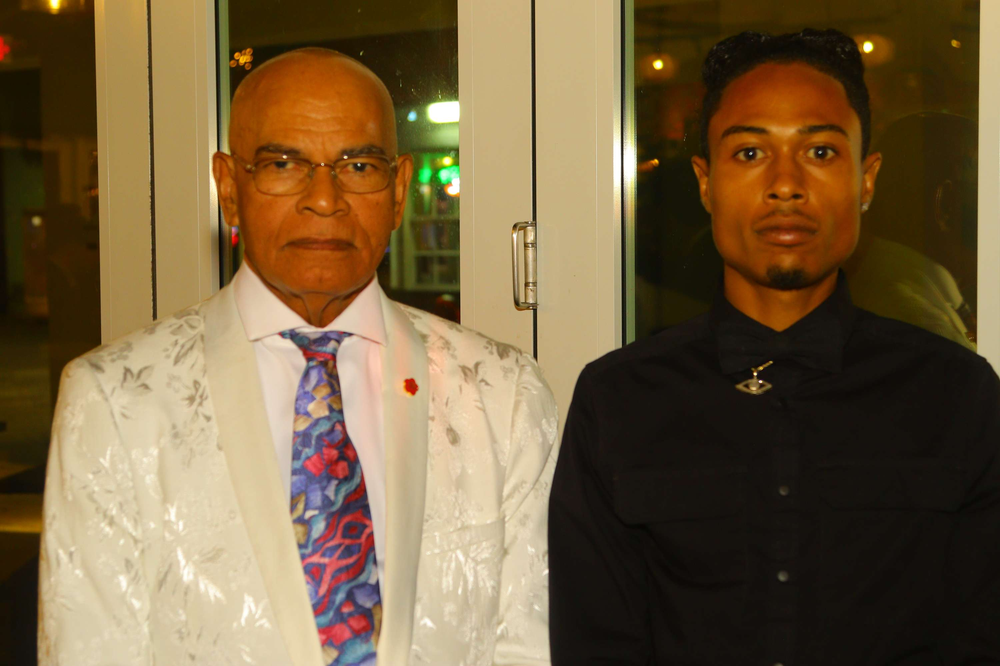 A man in a white suit and tie is standing next to a man in a black shirt.