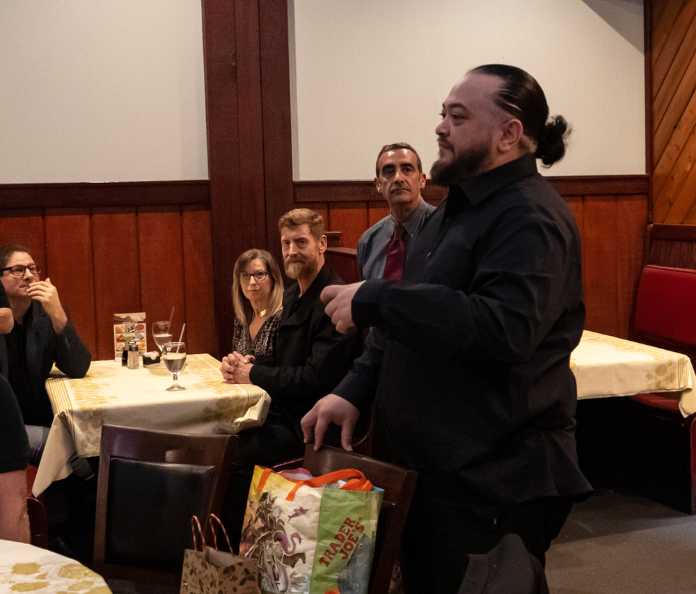 A man in a black shirt is standing in front of a group of people sitting at tables