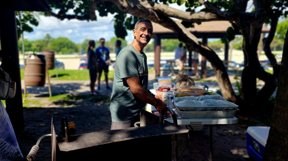 A man is standing in front of a grill in a park.