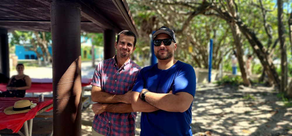 Two men are standing next to each other on a beach with their arms crossed.