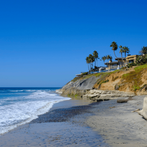 Beach with houses on a cliff, blue sky, ocean waves.