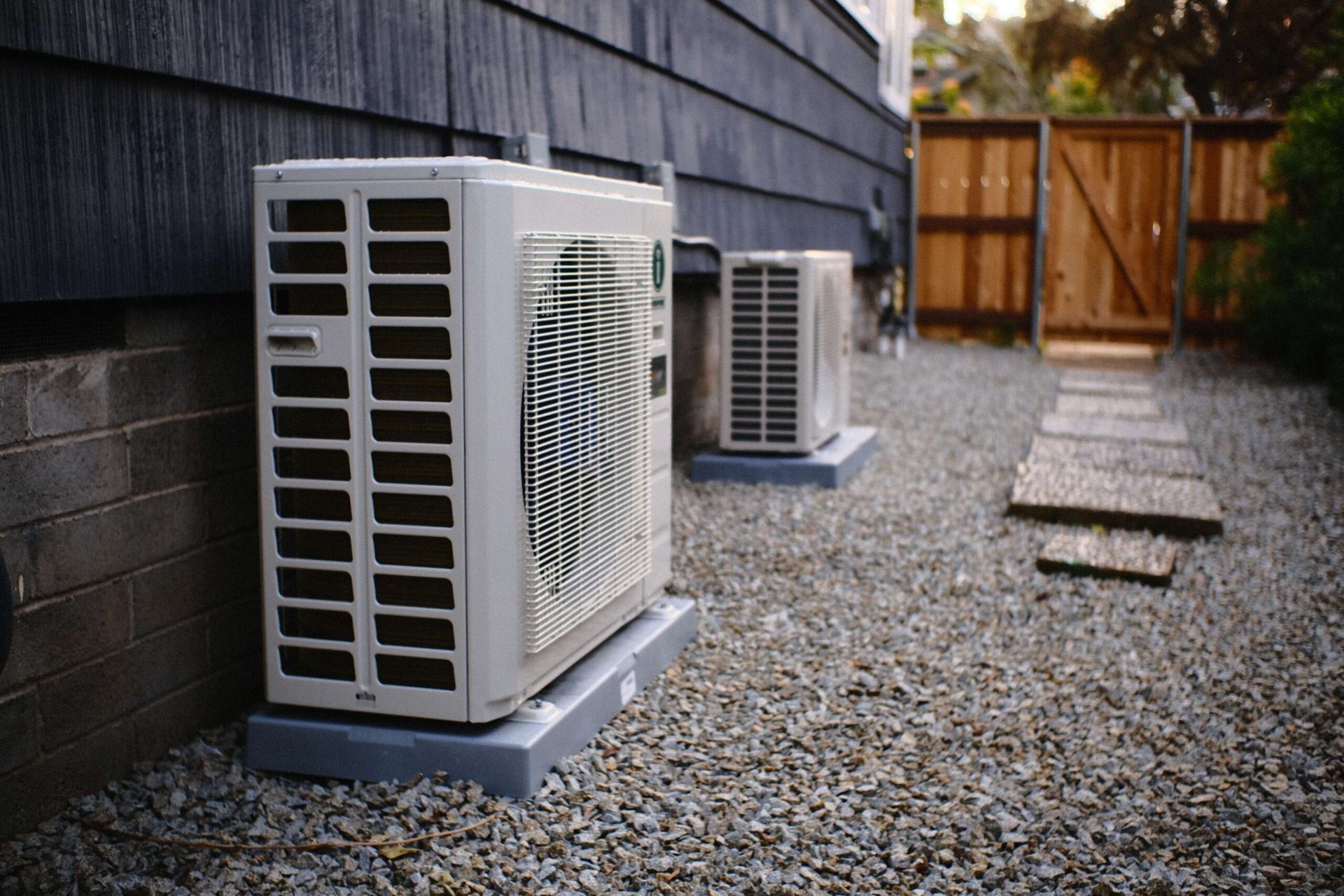 Two air conditioning units sit on concrete pads outside a dark-colored building, on a gravel bed with stepping stones.