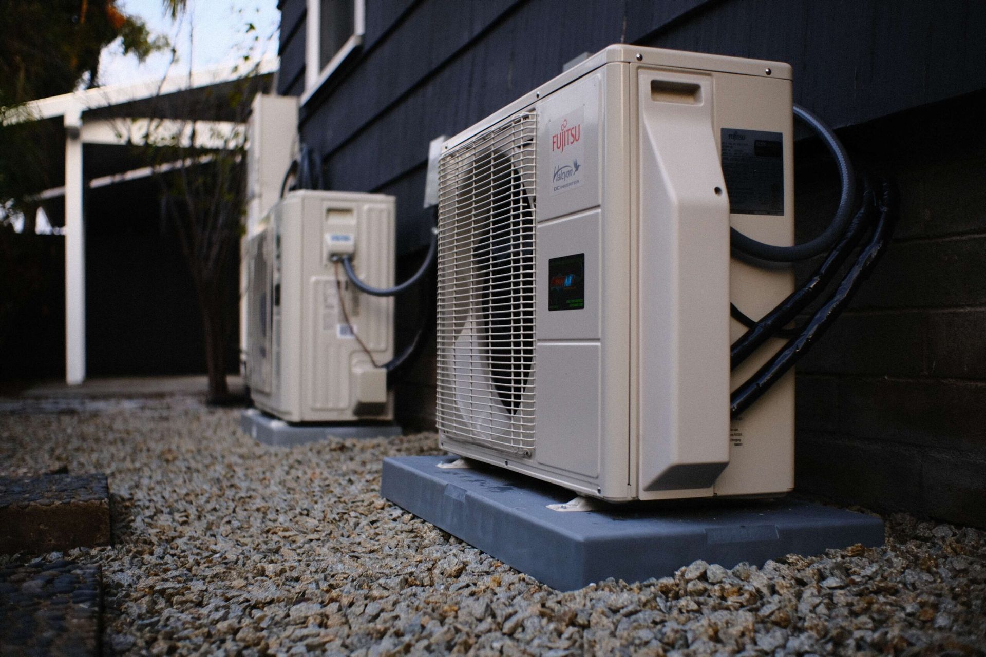 Two beige air conditioning units on concrete pads beside a dark house, surrounded by gravel.