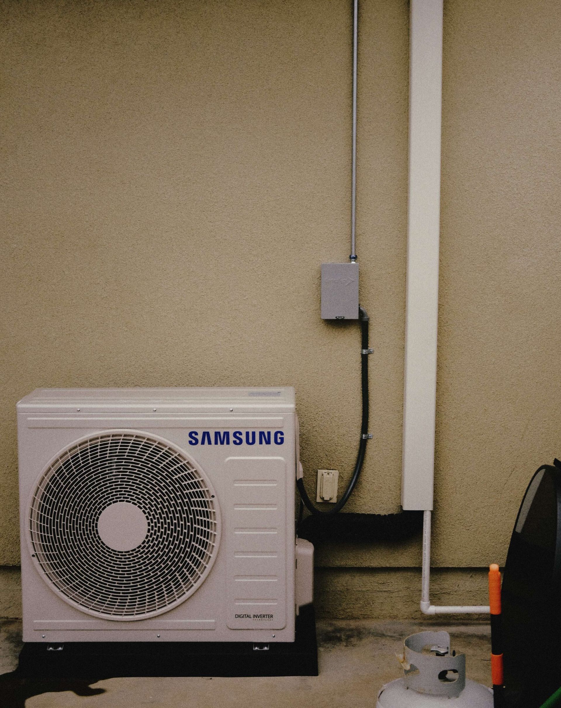 Samsung air conditioning unit against a light-colored wall, with electrical conduit and a propane tank.