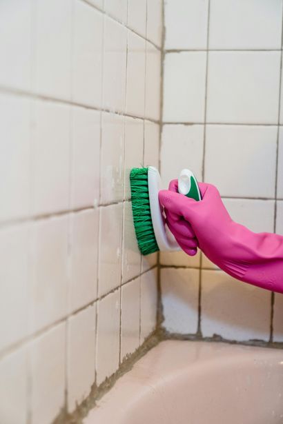 A hand in pink gloves scrubs white bathroom tiles with a green brush.