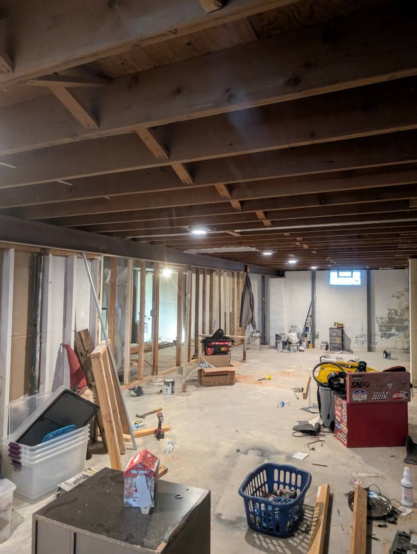 Unfinished basement with exposed ceiling joists, insulation, construction materials, and tools scattered on the floor.
