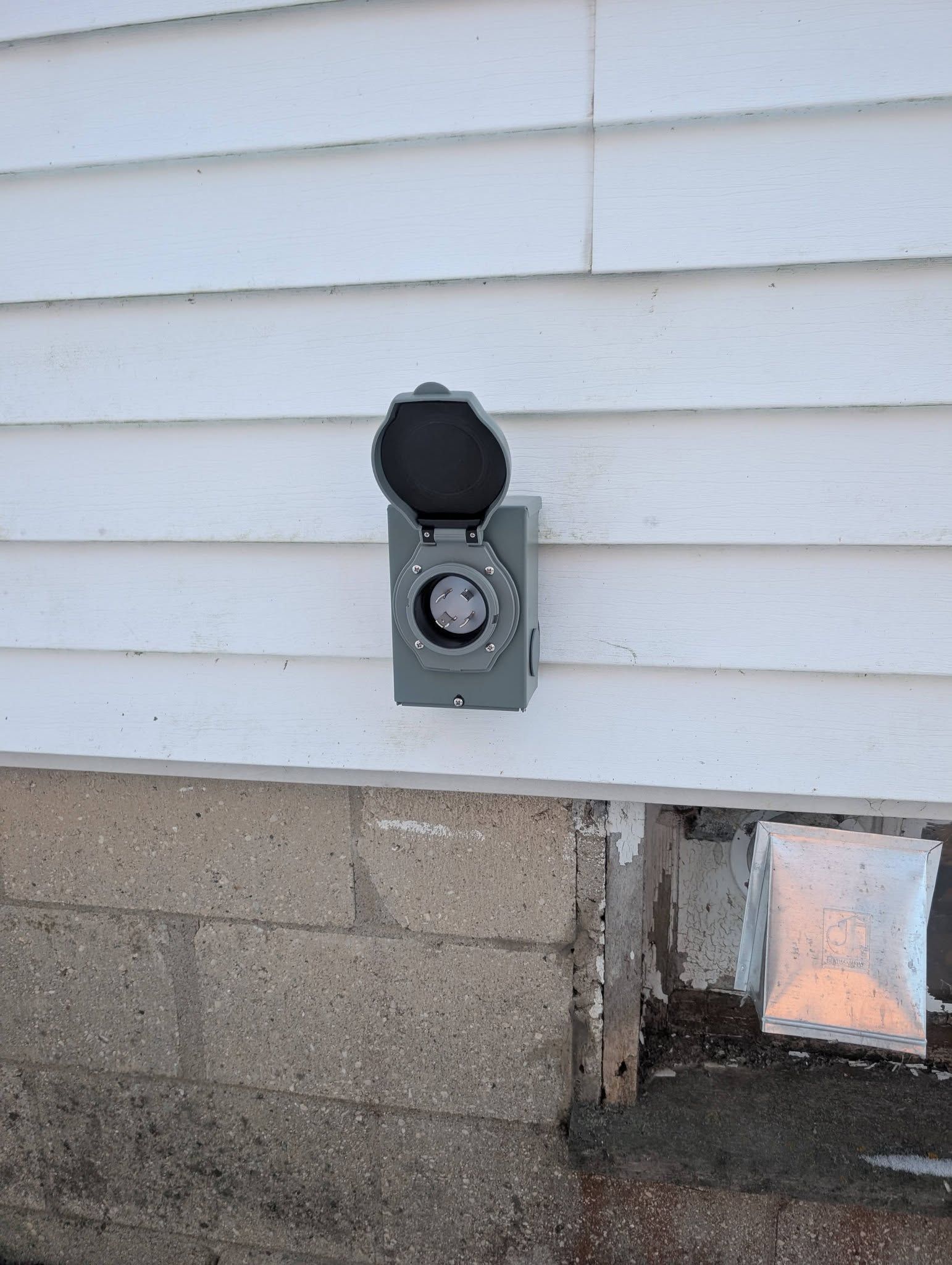 Exterior wall outlet with a black cover and a small vent opening on a house siding and cinder block wall