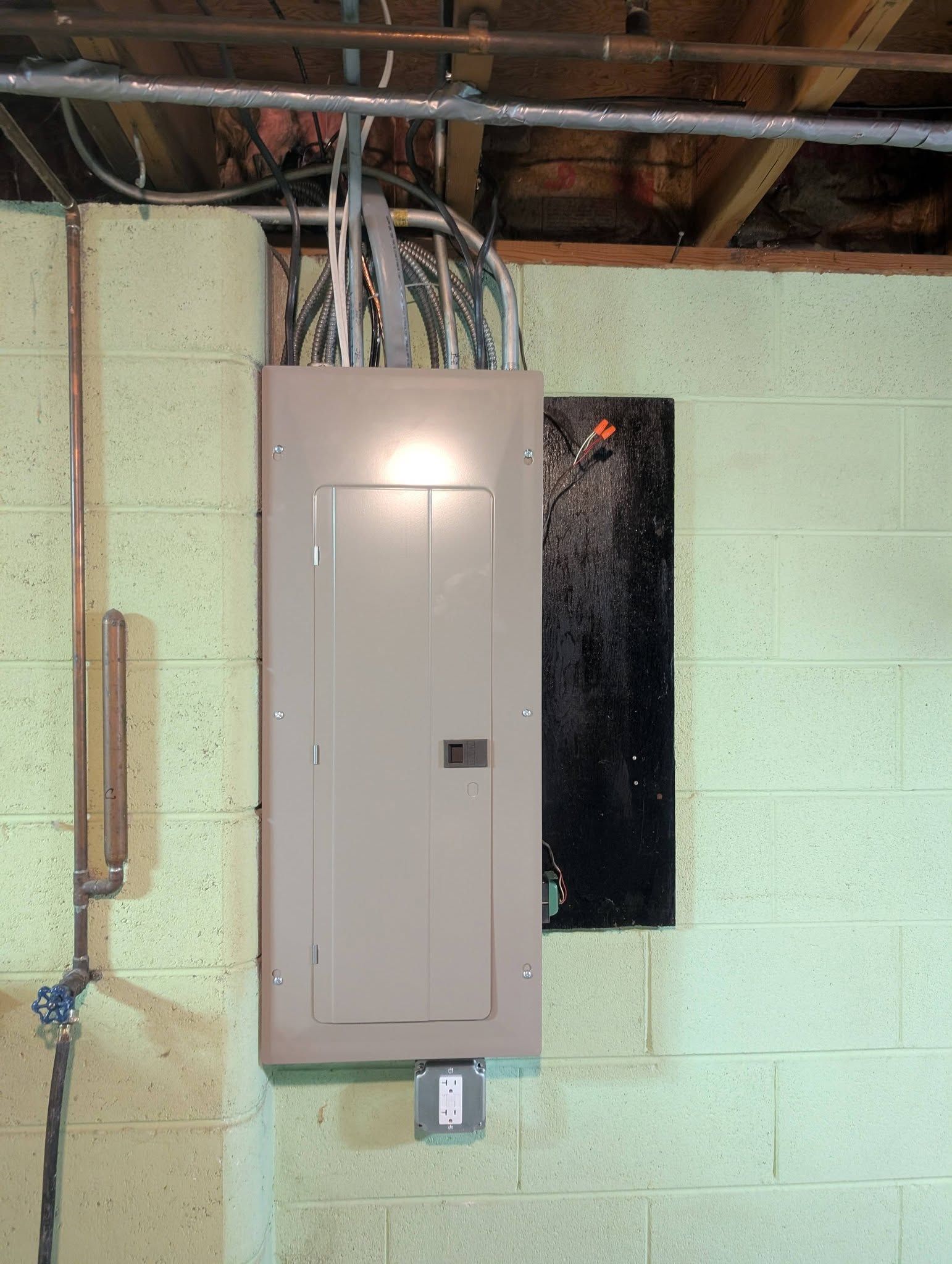 Open electrical breaker panel mounted on a cinder block basement wall, with exposed wiring above.