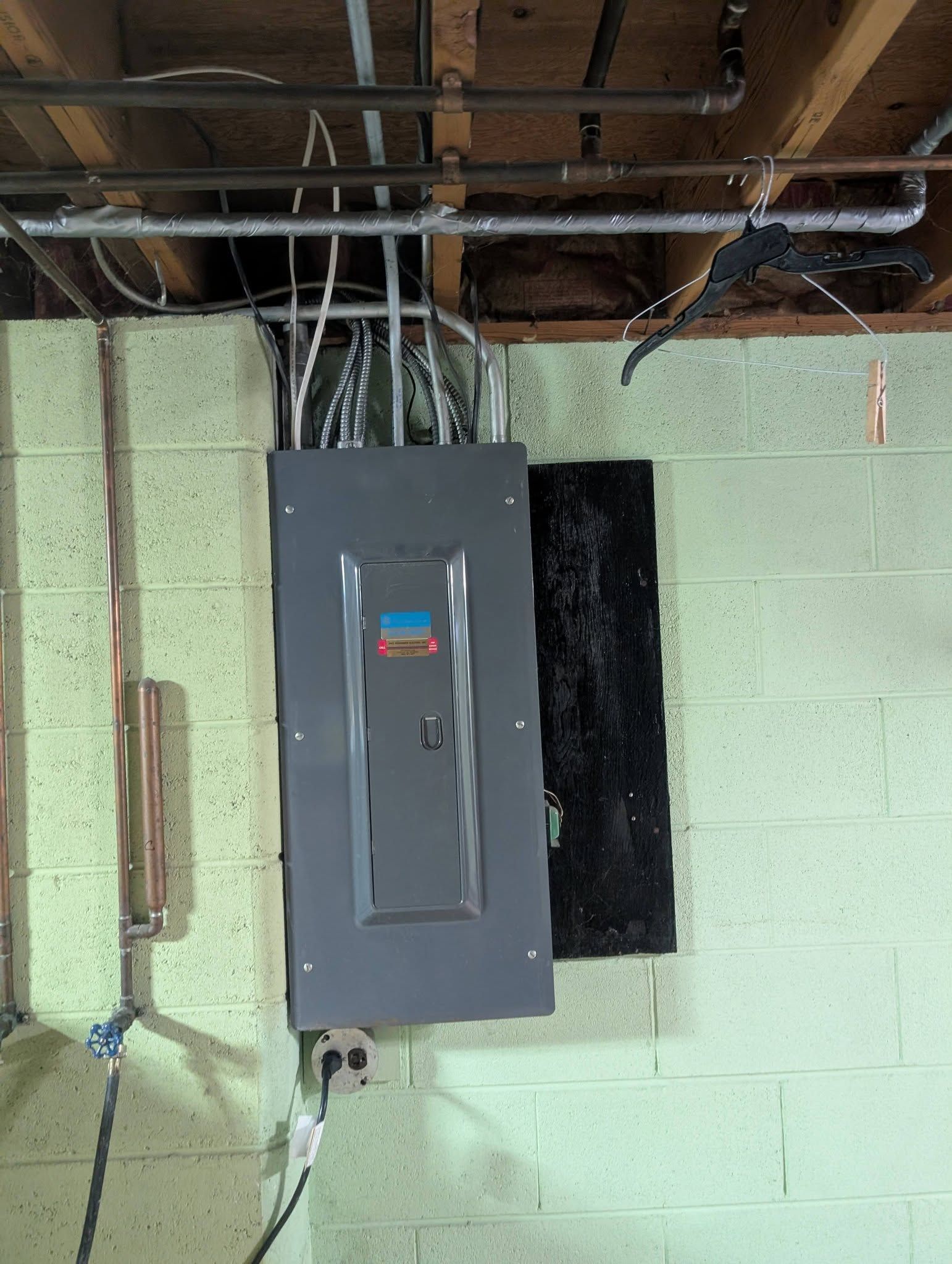 Gray electrical panel on a cinder block wall with exposed ceiling pipes and wiring nearby