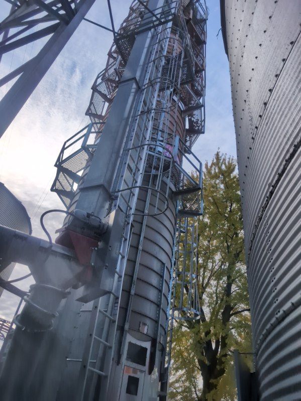 Tall metal silo with spiral stairs beside another silo, viewed from below against the sky and trees