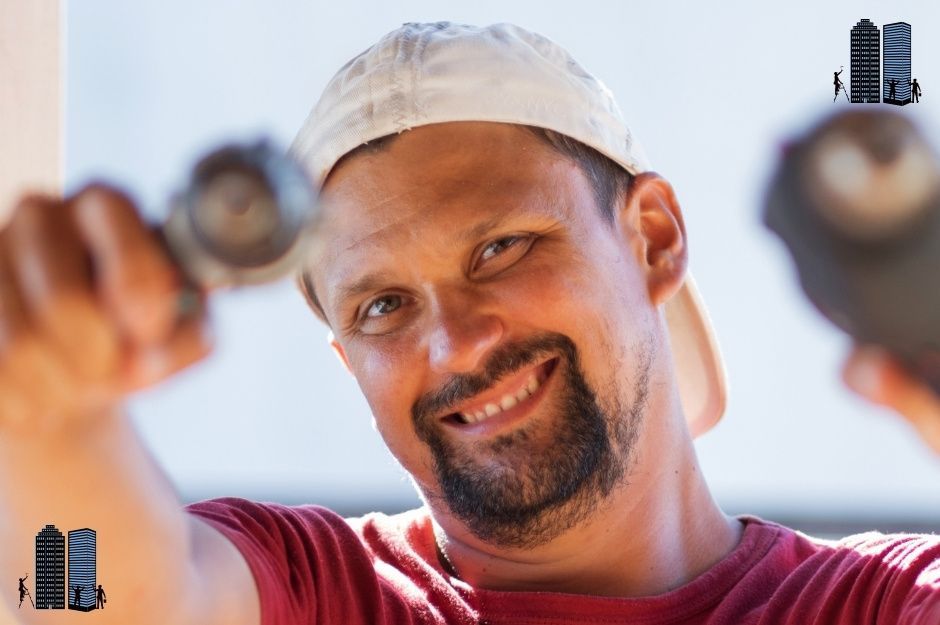 A close-up shot shows a smiling man, who appears to be a drywall contractor, wearing a white baseball cap and a red t-shirt, holding a power drill with a screw bit pointed towards the viewer, suggesting work related to drywall installation or repair in Brentwood, Burnaby, British Columbia.