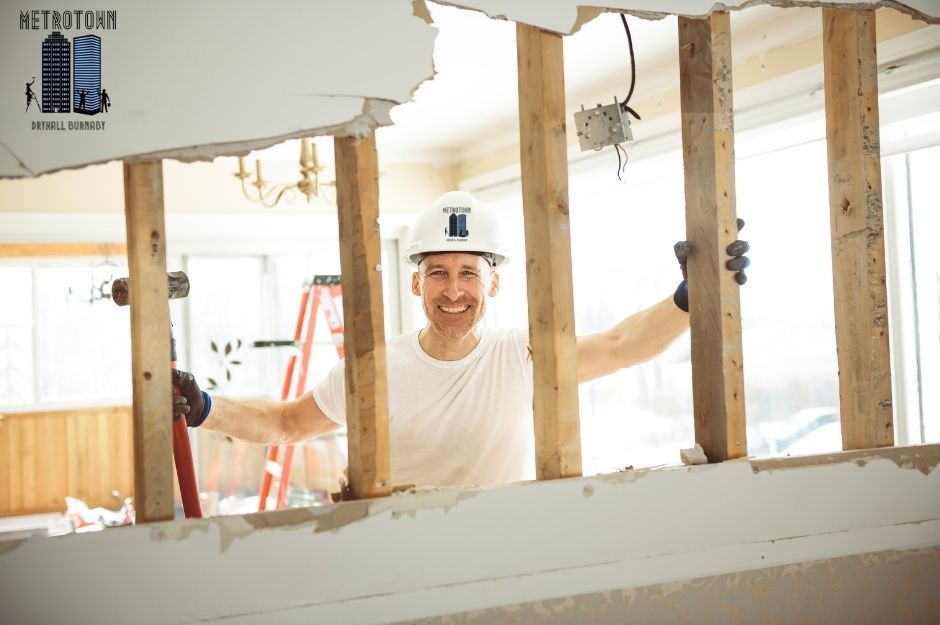 An expert from a drywall repair company in a white protective helmet is leaning on a wood panel while working on a residential drywall project. He positions himself near drywall panels in the home's construction zone with visible framing and wiring near 51a Ave, and 207 St in Langley, BC.