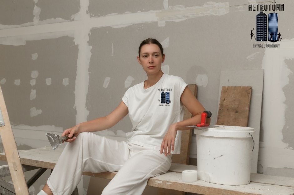 A drywall repair company worker sits on wooden planks at a commercial project site, wearing white work clothes. She holds a putty knife while taking a break, with a paint bucket, construction materials, and unfinished drywall visible around her near Blackburn Ave and 136 St, White Rock, BC.