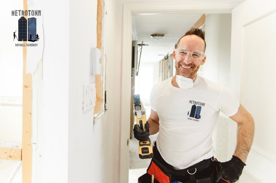 A drywall repair contractor in a white t-shirt smiles while holding a power drill, standing in a commercial hallway with white walls and wooden framing visible on Sherbrooke St, and Knox St in Sapperton, New Westminster, AZ.