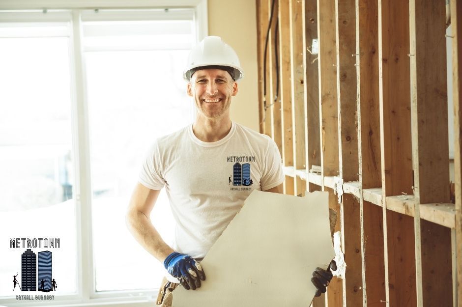 An experienced drywall repair contractor in a white t-shirt, a white hard hat, and blue gloves is cheerfully transporting drywall material during a commercial building renovation. He holds the large construction drywall panel securely while working in the framed commercial interior at Hammond Rd, and 193 St in Pitt Meadows, BC.