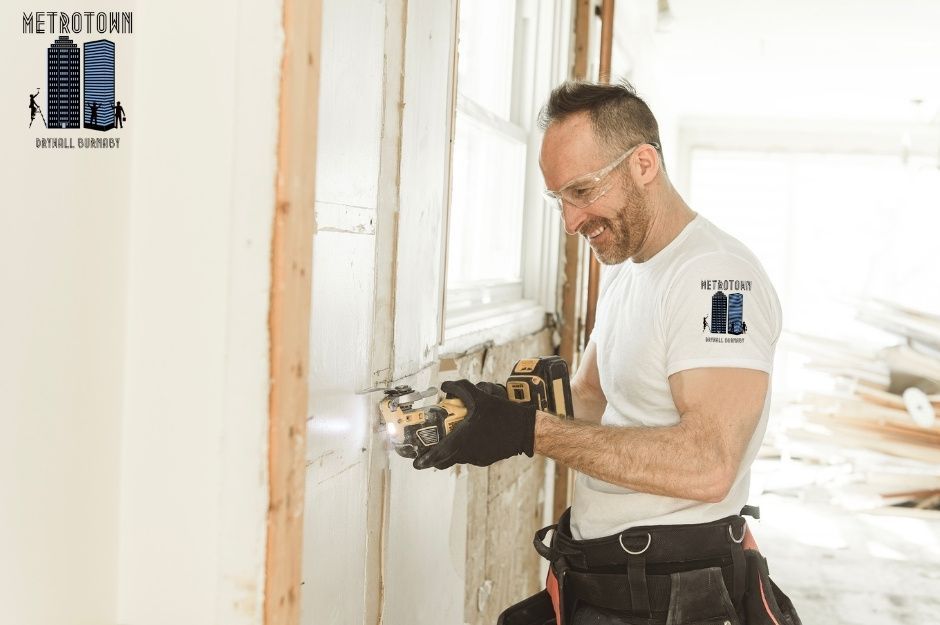 A professional drywall repair contractor in white t-shirt is using power tool to work on a commercial drywall installation. The commercial construction project is located near Kilmer St, and Prairie Ave in Port Coquitlam, BC.