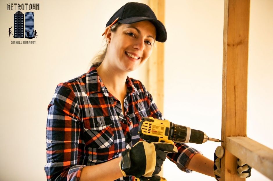 A drywall repair contractor in a black cap and plaid shirt smiles while holding a power drill, working on plasterboard repair in a room with white walls and wood trim on Eyremount Dr and Fairmile Rd, West Vancouver, AZ.