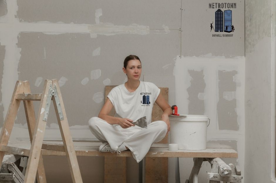 A drywall repair contractor sits on wooden planks during a residential renovation, wearing white work clothes. She holds a paintbrush while taking a break, with a stepladder, paint bucket, and construction materials visible around the unfinished room at 201 St, and 70a Ave in Willoughby, BC.