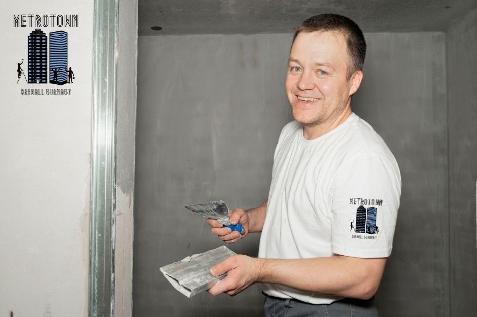 A drywall repair handyman smiles while holding a putty knife and hawk during a residential project. He wears a white shirt and stands near freshly applied drywall compound on the wall near Lucas Rd, and Lismer Gate in Richmond, BC.