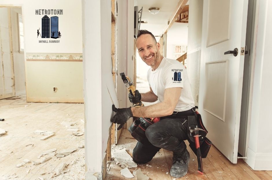 A drywall repair specialist in a white t-shirt kneels while holding a power drill, working for a residential project with white walls and debris on the floor on Cecile Pl, and Cecile Dr in Port Moody, AZ.
