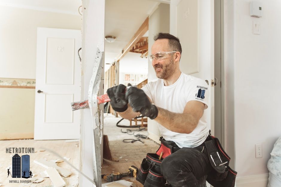 A smiling drywall repair technician in a white t-shirt and dark pants wearing safety glasses sits while holding a power tool, working on a residential sheetrock project with white walls and construction materials near Galloway Ave, and Soball St in Burke Mountain, Coquitlam, AZ.