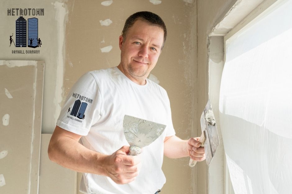 A drywall repair technician applies joint compound using a putty knife during a commercial project. He wears a white shirt and smiles while working on the drywall seams, with patch repairs near Delta St, and 48 Ave in Delta, BC.