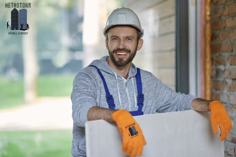 An experienced drywall repair technician wearing a distinctive orange work gloves and casual construction attire is smiling while working in residential drywall completion. The professional stands against his quality installation work in a bright home interior in Dewdney Trunk Rd & 256 St., Maple Ridge, BC.