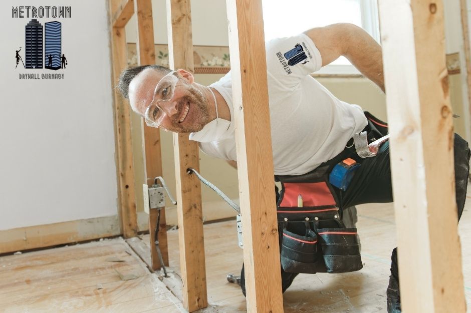 A drywall repair technician, wearing a white T-shirt, kneels while using a power drill among wooden framing studs doing a residential sheetrock project near Lonsdale Avenue and West 21st Street in Central Lonsdale, North Vancouver, AZ.