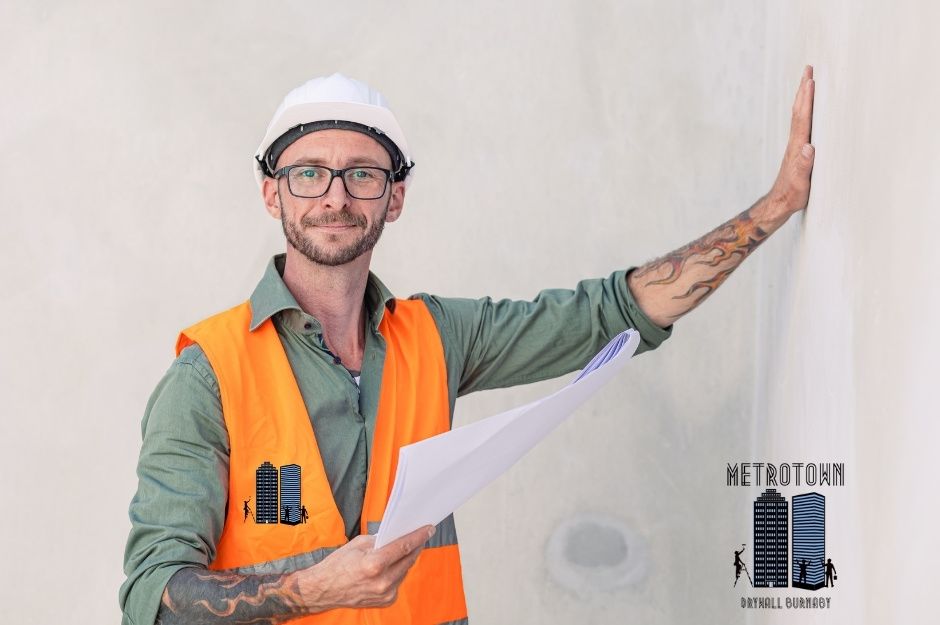 A drywall repair technician wearing a white hard hat, glasses, an orange safety vest over a green shirt, and holding paperwork. A smiling man stands against a white drywall background in a commercial construction site near Charles St, and Renfrew St, Vancouver, BC.