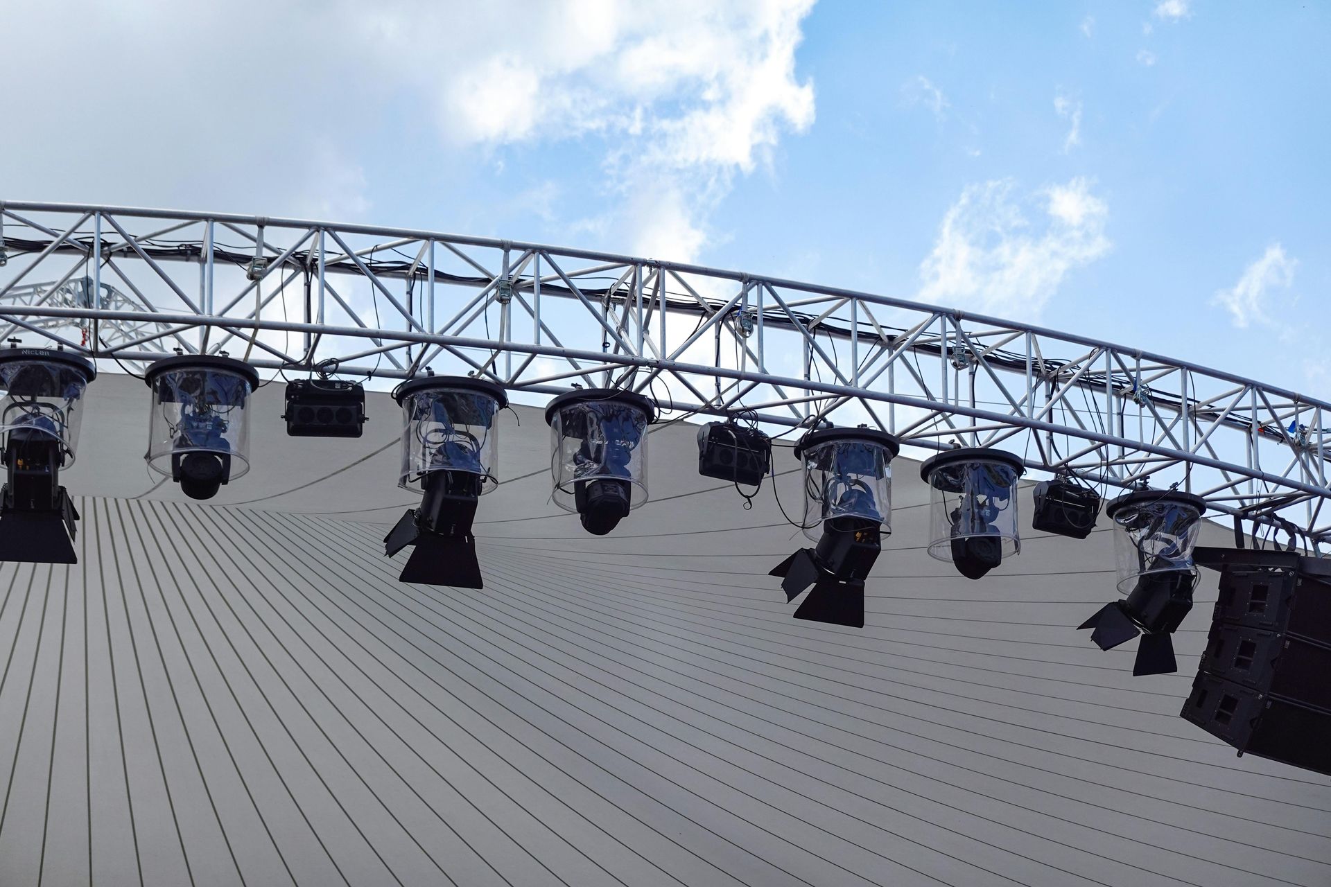 Stage lights hanging from a metal truss under a cloudy sky.