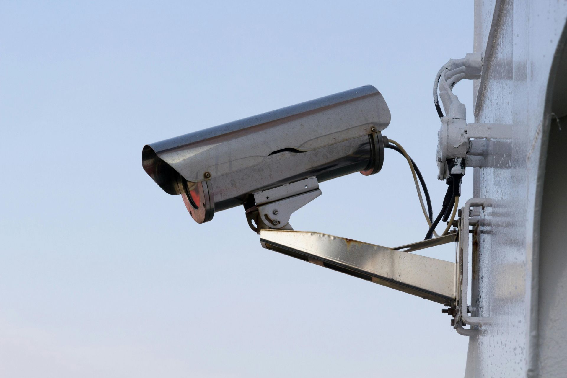Security camera, silver and black, mounted on a white pole, against a light blue sky.