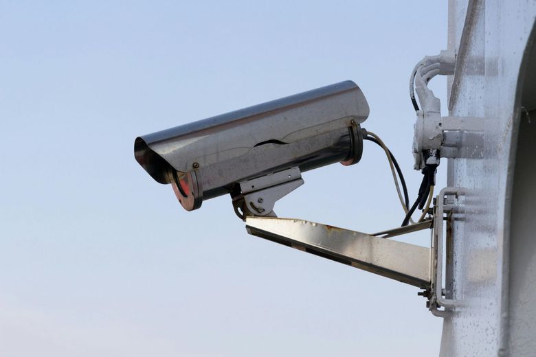 Security camera, silver and black, mounted on a white pole, against a light blue sky.