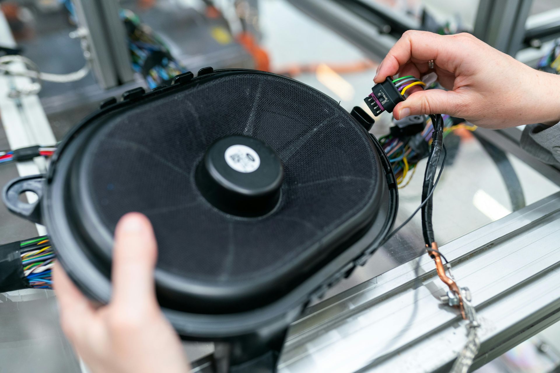 Hands connecting a wire harness to a car speaker on an assembly line.