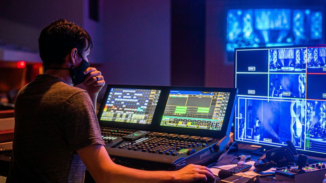 Man in mask operating a lighting console with screens, controlling a stage performance, dimly lit interior.