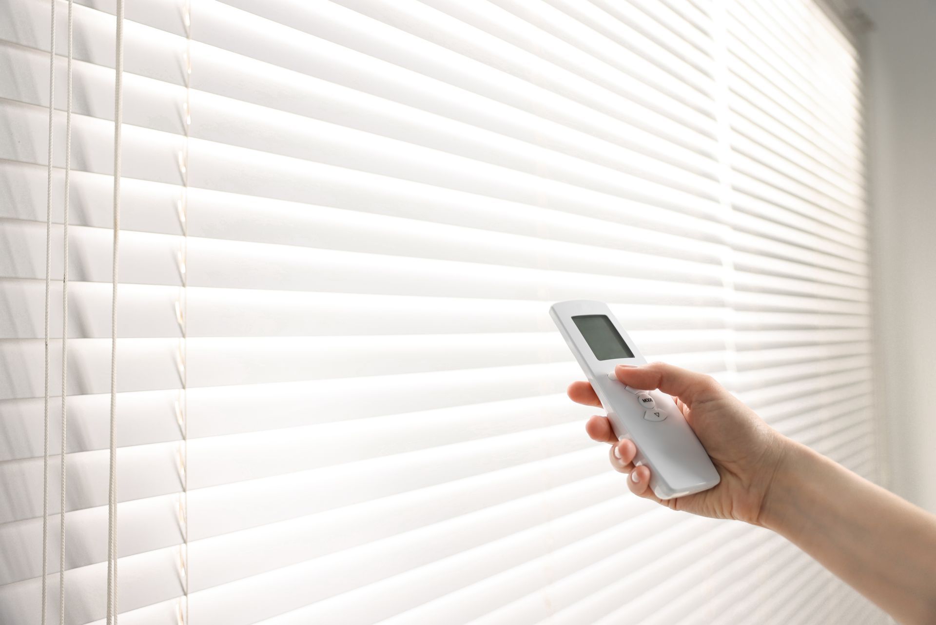 Hand holding remote, adjusting white window blinds in a brightly lit room.