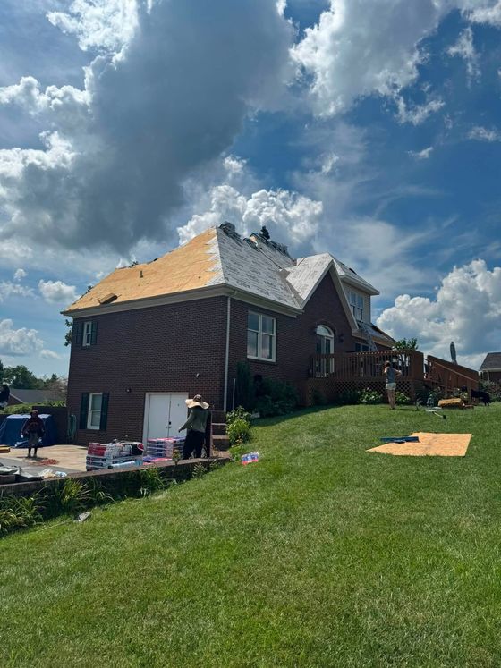 Brick house under repair with exposed roof framing, tools on the lawn, and dramatic cloudy sky