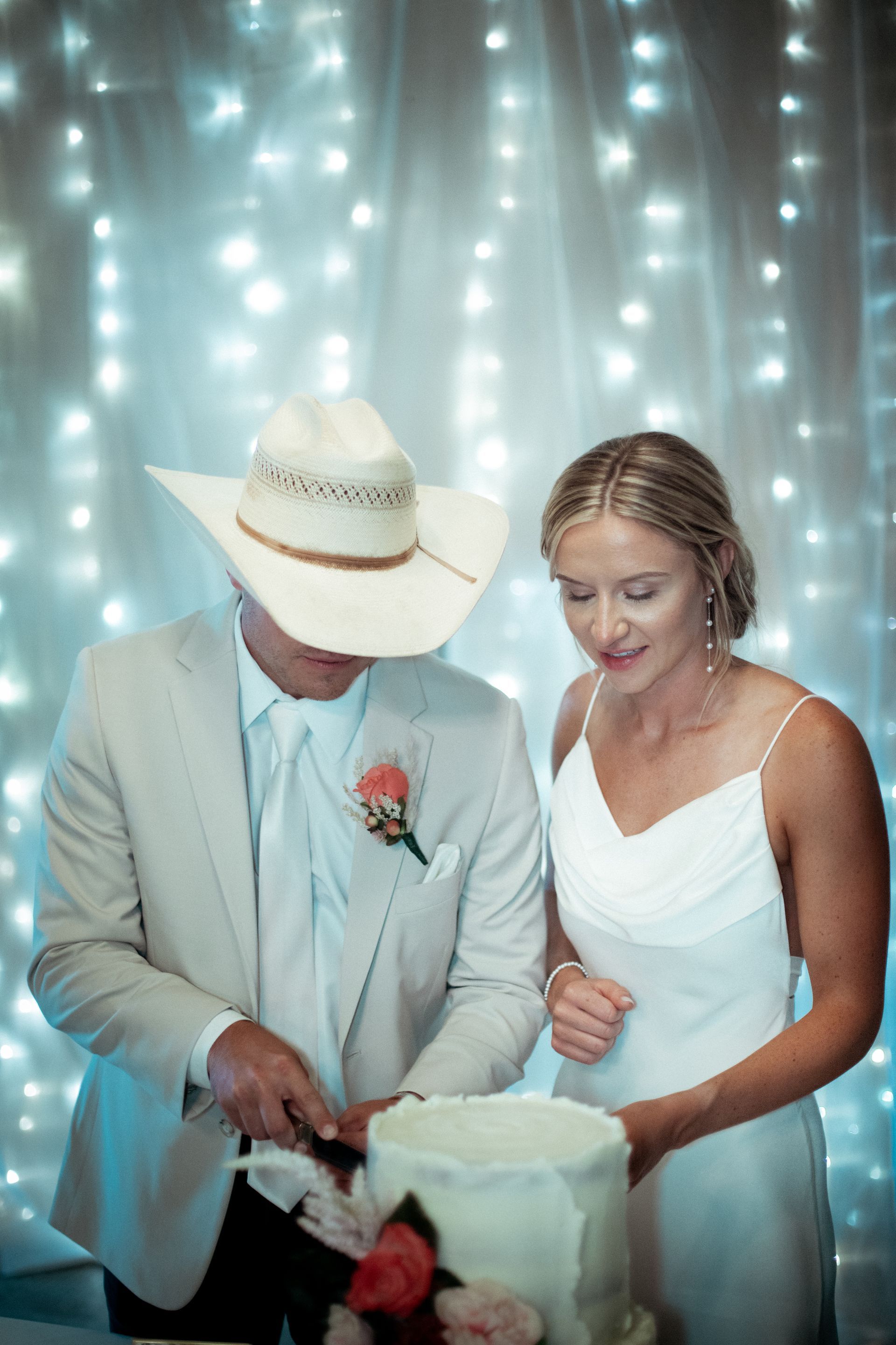 A bride and groom are cutting their wedding cake . the bride is wearing a cowboy hat.